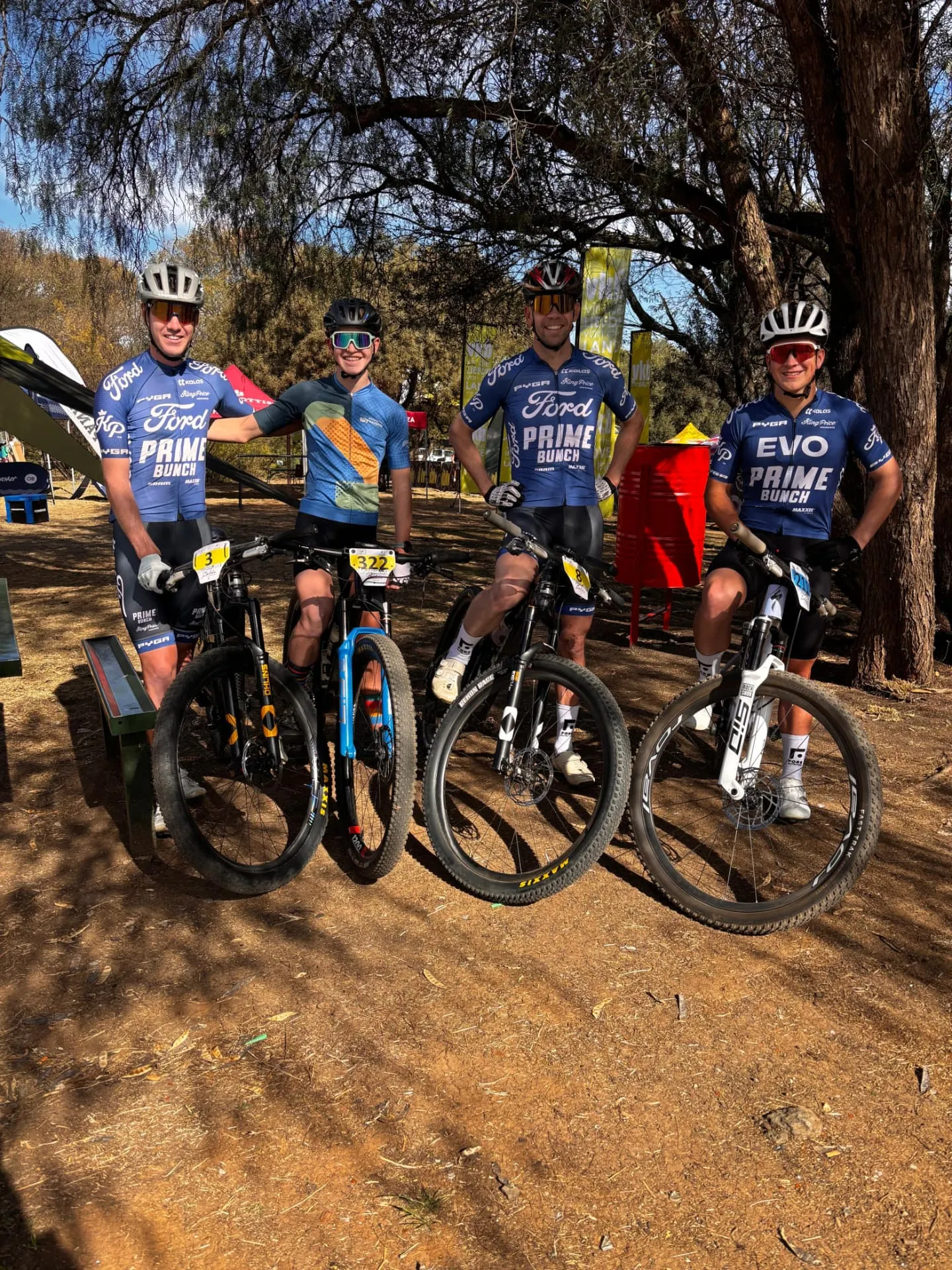 Four Prime Bunch EVO academy riders in blue kit standing with their mountain bikes under trees