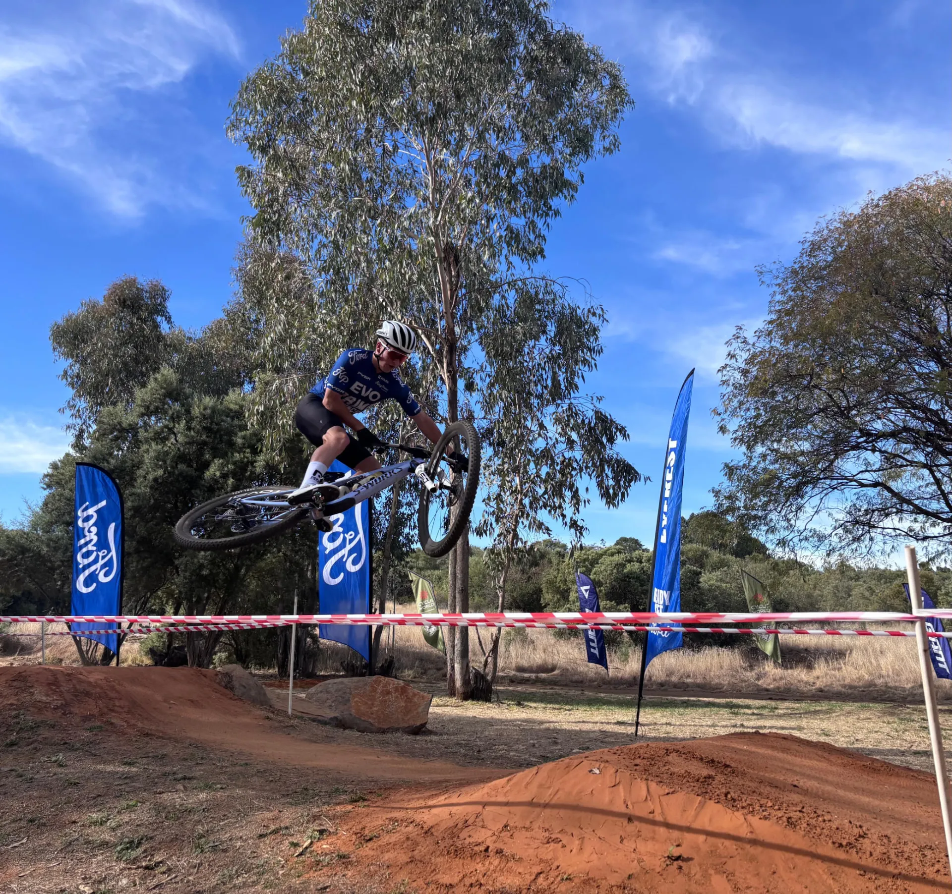 Academy rider in blue EVO Prime Bunch kit jumping a mountain bike over a dirt feature